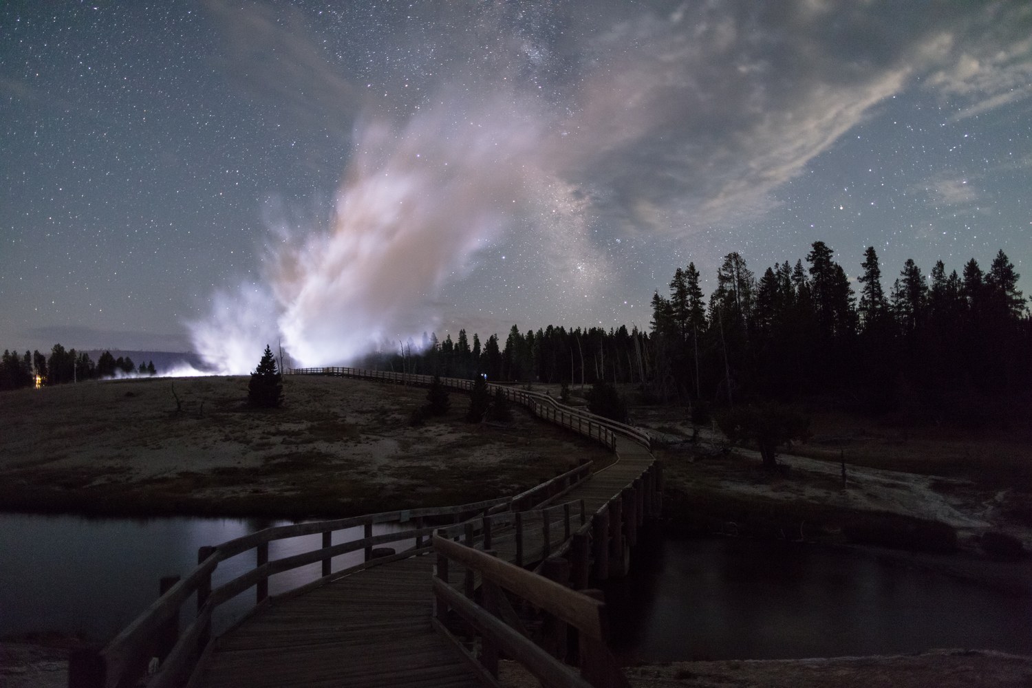 Castle Geyser erupting at night beside an empty boardwalk under a starry sky.